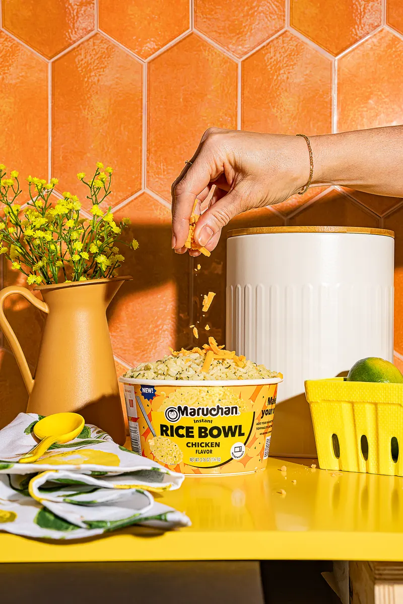 A hand sprinkling shredded cheese over a Maruchan Chicken Flavor Rice Bowl